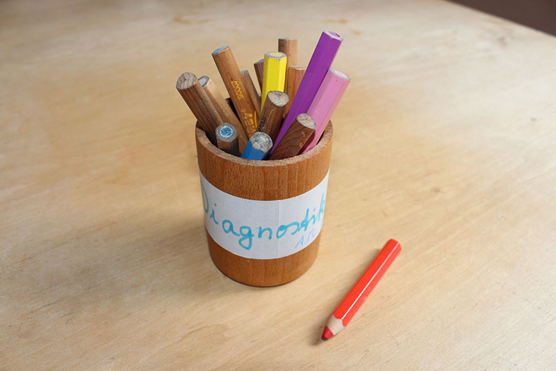 Ein Holzbecher mit farbigen Wachsmalstiften steht auf einem Tisch
